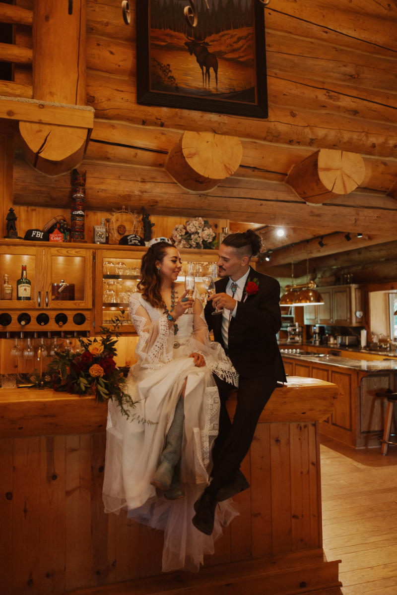 Bride and groom sit on top of wine bar drinking champagne at their New Mexico mountain wedding venues bar.