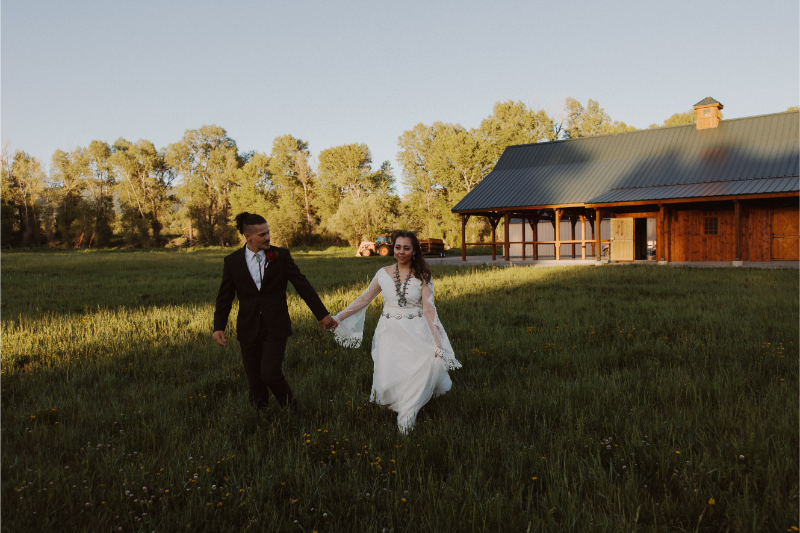 Bride and groom walk in grass field with views their New Mexico mountain wedding venues behind them.