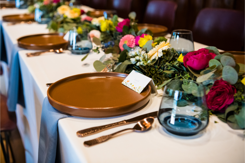 Wedding reception table setting for New Mexico celebration with gold silverware, blue glassware, and long, low wildflower centerpiece lining the long rectangle table.