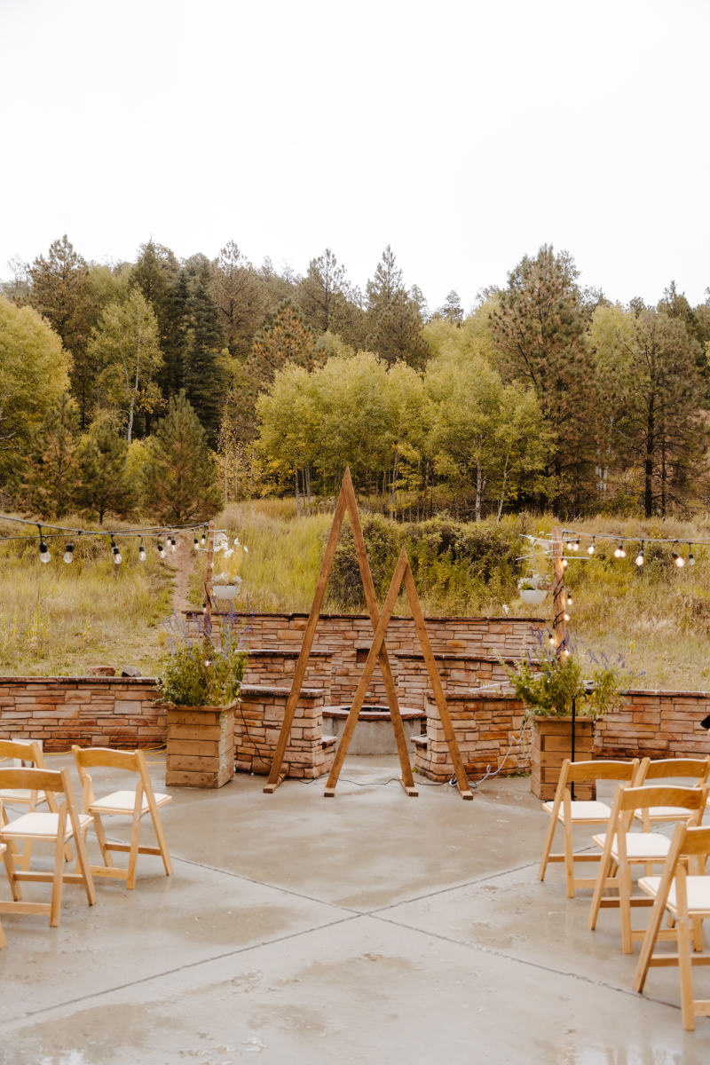 An outdoor new mexico micro wedding ceremony setup on a concrete patio, featuring two crossed wooden A-frame arches as the altar, flanked by stacked stone planters with wildflowers and greenery. String lights drape across the space and natural wooden folding chairs line both sides of the aisle, with a backdrop of golden aspens and pine trees in early fall.