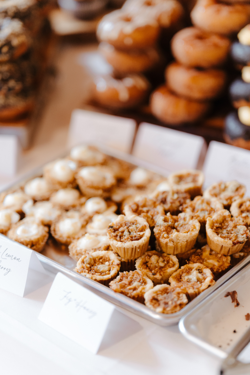 A styled dessert table spread at an intimate wedding reception featuring mini tart bites labeled with handwritten calligraphy signs reading "Lemon Berry" and "Fig Honey," arranged in silver trays alongside stacked donuts in the blurred background.