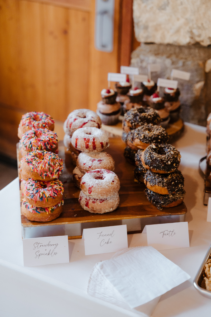A fun and festive donut display on a rustic wooden board at a new mexico micro wedding reception, with stacked rows of donuts in three flavors: Strawberry Sprinkles, Funnel Cake, and Turtle, each labeled with handwritten calligraphy place cards. Cupcakes with raspberry toppers are staged behind them.