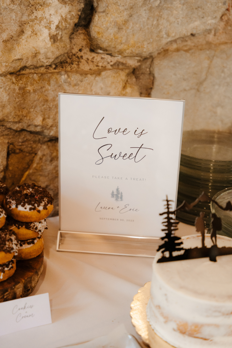 "Love is Sweet" acrylic dessert table sign for Laura and Eric's September 30, 2023 wedding, displayed against a stone wall backdrop beside a white naked cake topped with a mountain silhouette cake topper and a tower of cookies and cream donuts.
