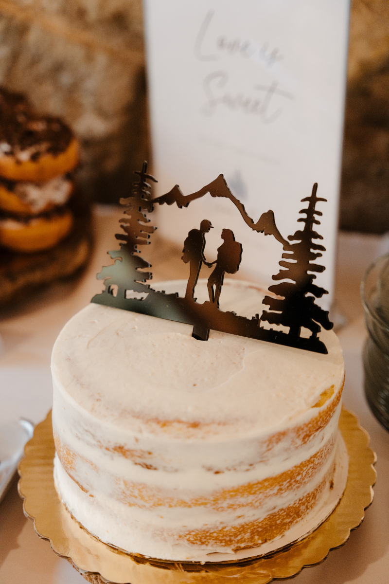 A semi-naked white wedding cake topped with a laser-cut black acrylic cake topper featuring two hikers standing hand-in-hand amid pine trees and mountain peaks, reflecting the couple's adventurous spirit. The "Love is Sweet" sign is softly visible in the background.