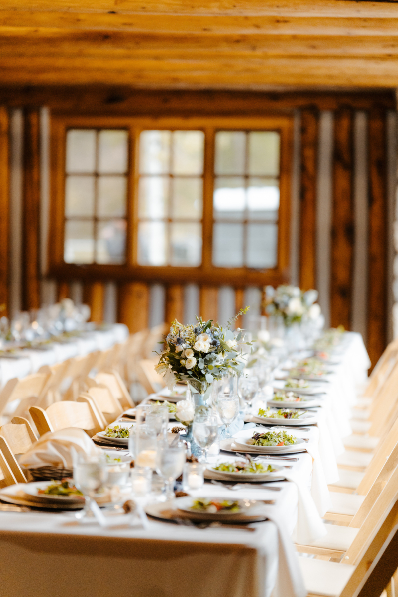 A sunlit banquet table fully set for the wedding reception inside a mountain log cabin venue, with dressed salads already plated on wood charger plates, crystal stemware glinting in natural window light, and low floral centerpieces of white roses, dusty miller, and eucalyptus along a soft blue runner.