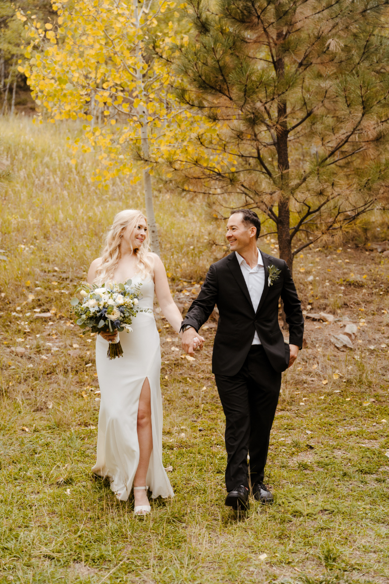 The bride and groom walk hand-in-hand through a mountain meadow surrounded by golden aspen and tall pine trees, both smiling at each other. The bride carries a lush bouquet of white roses, blue hydrangea, and eucalyptus, and the groom's thistle boutonniere complements the fall palette in this new mexico micro wedding portrait.