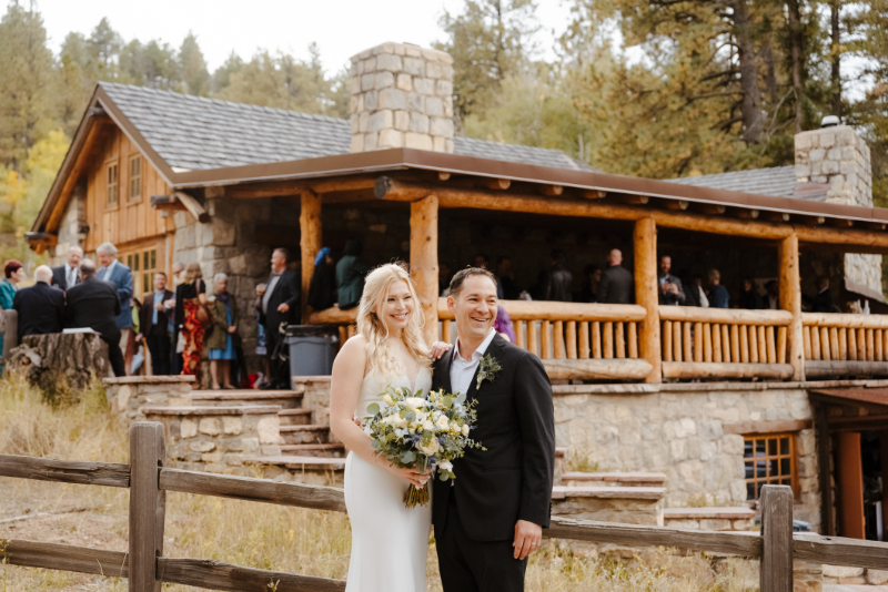 Bride and groom poses together in front of a stunning log and stone mountain lodge venue following their New Mexico micro wedding ceremony, guests mingling on the covered porch behind them. The bride holds her garden bouquet as they both beam at the camera, surrounded by the wild grasses and pine forest of the New Mexico mountains.