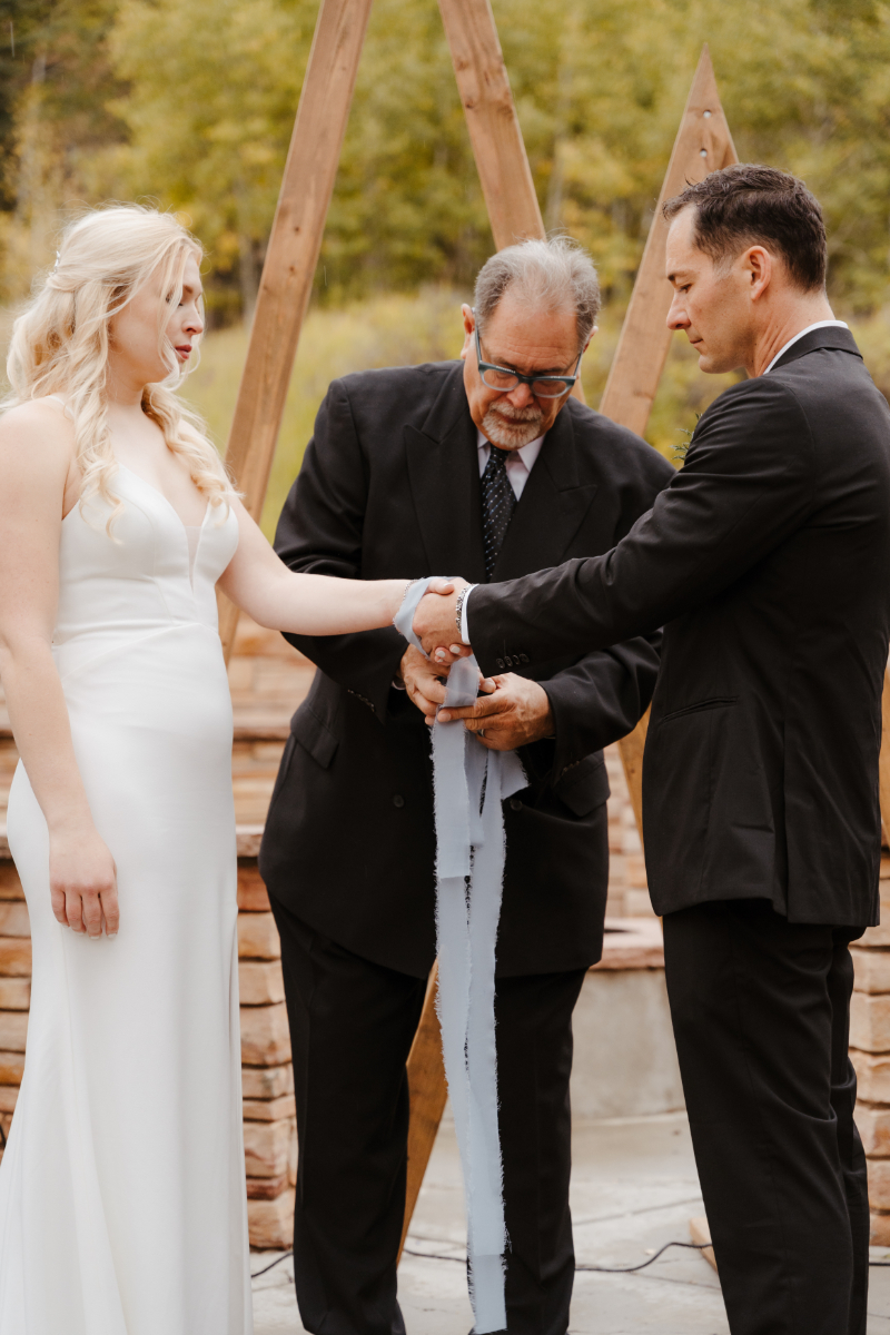 A wedding officiant performs a handfasting ceremony at a new mexico micro wedding, wrapping a pale blue ribbon around the joined hands of the bride and groom. The bride wears a simple white slip gown while the groom is dressed in a dark suit, both standing before rustic wooden arch structures.
