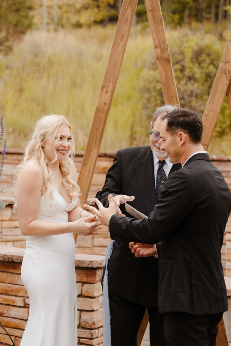 A joyful ring exchange moment at an intimate new mexico micro wedding ceremony. The bride laughs brightly as the groom slides her wedding band onto her finger, with the smiling officiant watching behind them and the crossed wooden arch and fall foliage filling the background.