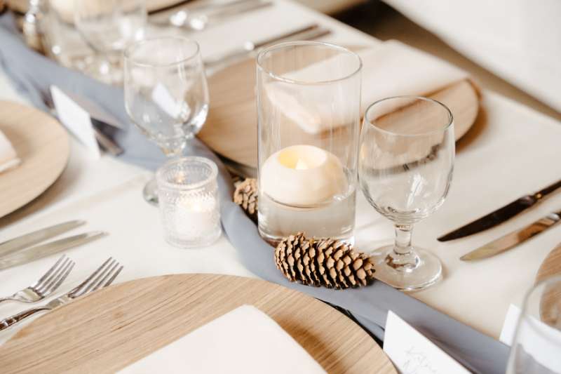 A top-down view of an elegant yet natural wedding place setting with a circular wood charger plate, a neatly folded ivory linen napkin, silver flatware, and crystal stemware. Pinecones and votive candles are nestled along the dusty blue table runner, with handwritten place cards visible in the background.
