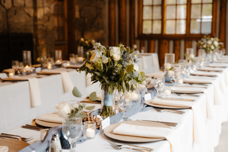 A wide shot of the reception dining room at a new mexico micro wedding, showing the full length of a long white-clothed table with wood charger plates, crystal goblets, ivory napkins, dusty blue runners, and low floral arrangements of white blooms and greenery beneath a ceiling of warm pine logs and large windows.