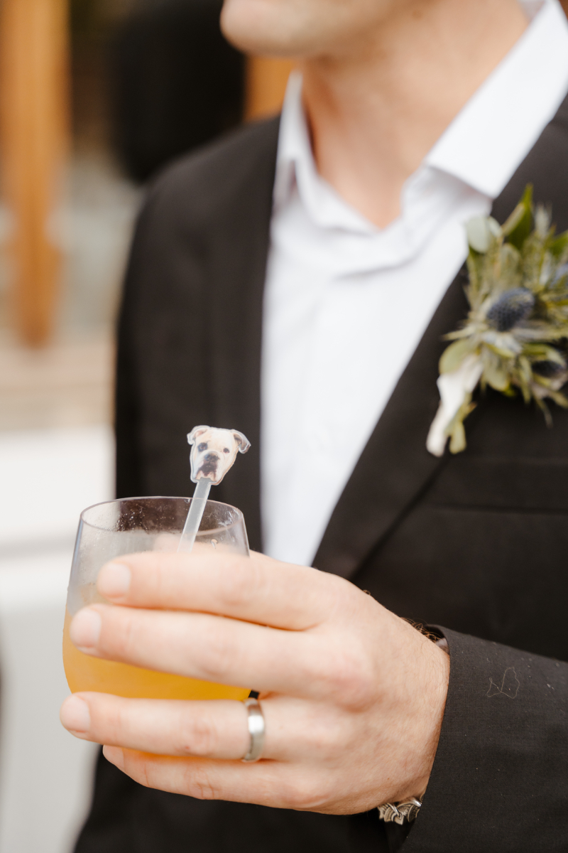 A detail shot of the groom holding a cocktail hour drink featuring a custom dog-face straw topper shaped like a white bulldog, nodding to the couple's beloved pets. His boutonniere of blue thistle and eucalyptus is visible on his lapel, and a wedding band is on his hand.