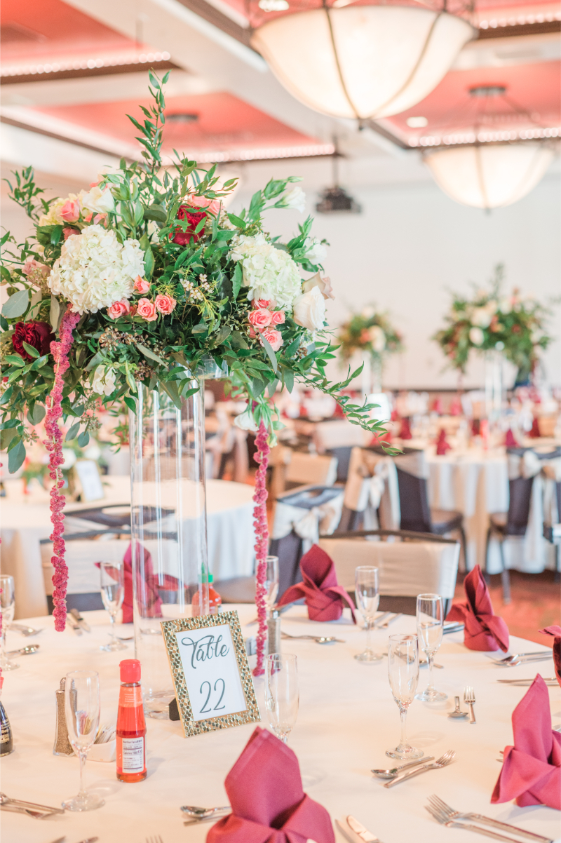 Tall clear glass centerpiece with lush white hydrangea, pink roses, and cascading burgundy amaranthus above a round table setting.