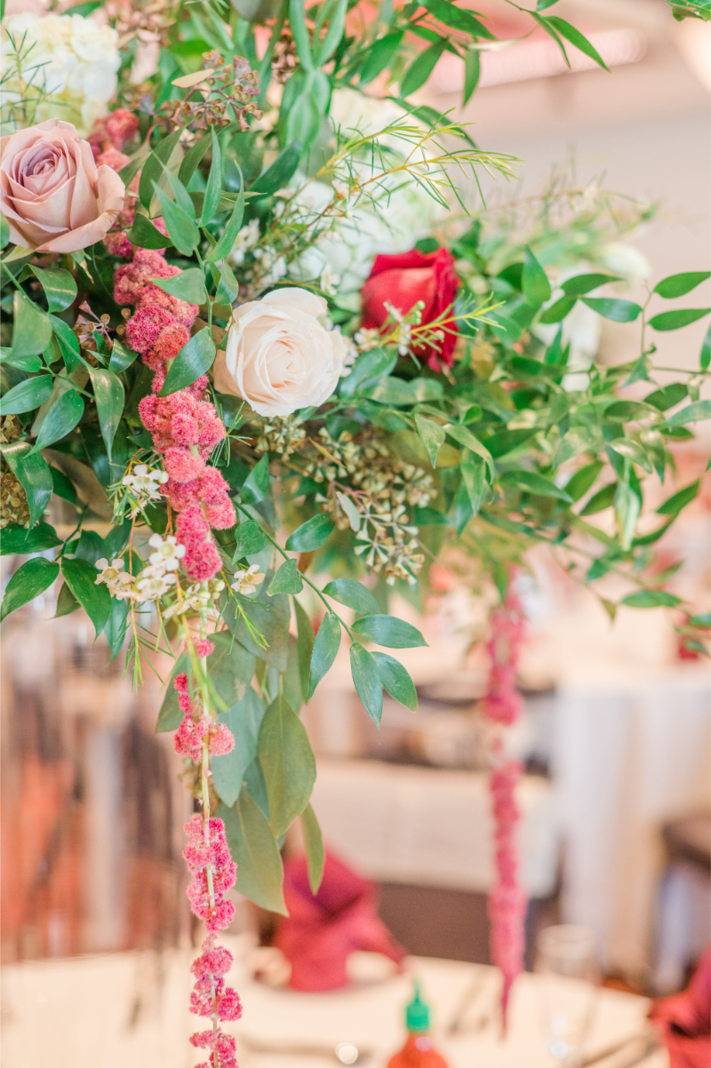 Close-up of a tall floral centerpiece with white roses, blush blooms, and trailing pink amaranthus against lush greenery.
