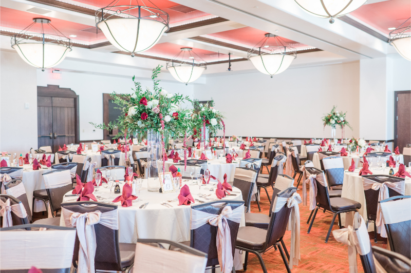 Spacious ballroom reception with tall flower arrangements in glass vases on every round table, accented by deep red napkins.