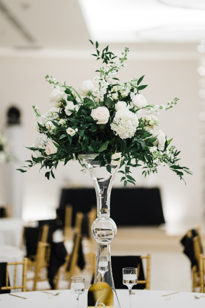 Tall flower arrangements with white roses, hydrangea, and wild greenery in a clear glass trumpet vase against a black and white ballroom setting.