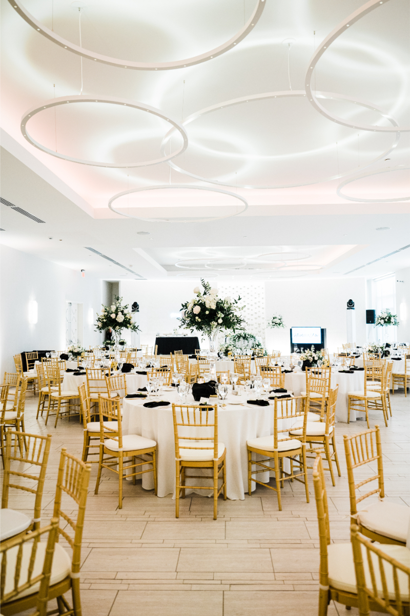 Full ballroom view with tall white and green floral centerpieces on every table, set against a modern white venue with sculptural ceiling detail.