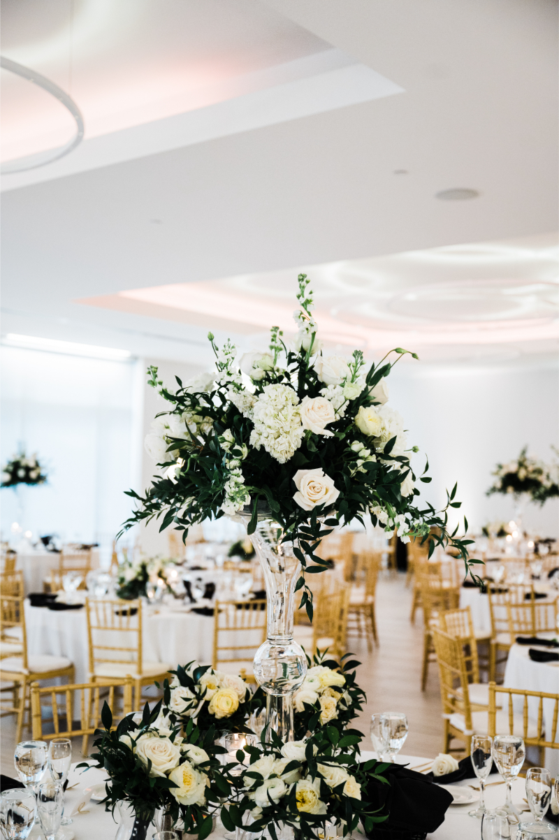 Full ballroom view with tall white and green floral centerpieces on every table, set against a modern white venue with sculptural ceiling detail