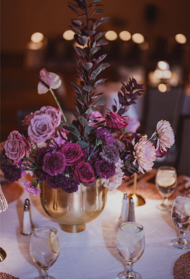 Low gold bowl centerpiece with layered jewel-toned blooms including mauve roses, magenta carnations, and deep burgundy florals.