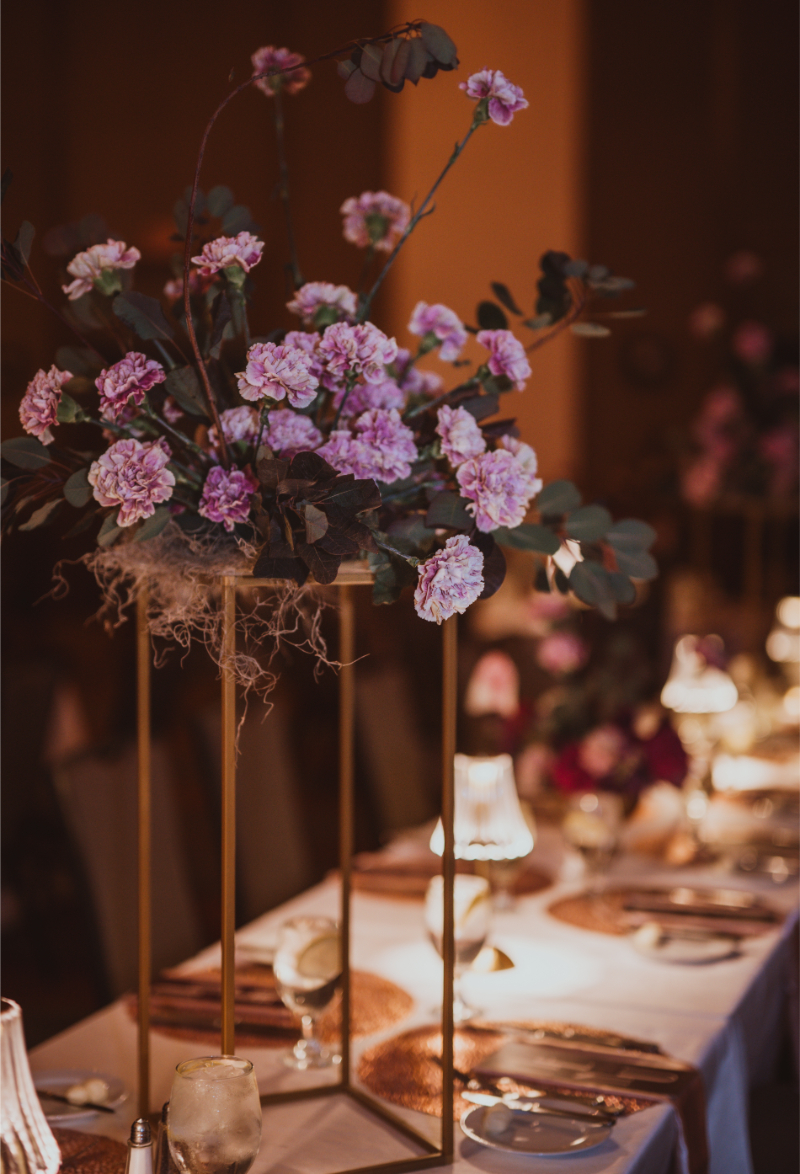 Tall flower arrangements with lavender carnations and dark eucalyptus on gold geometric stands along a white reception table.