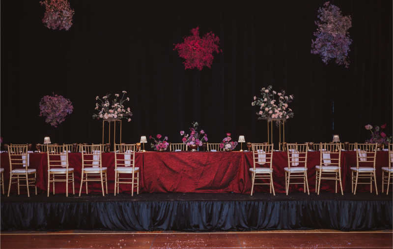 Elevated floral installations suspended above a dramatic head table with deep red linens and gold chiavari chairs.