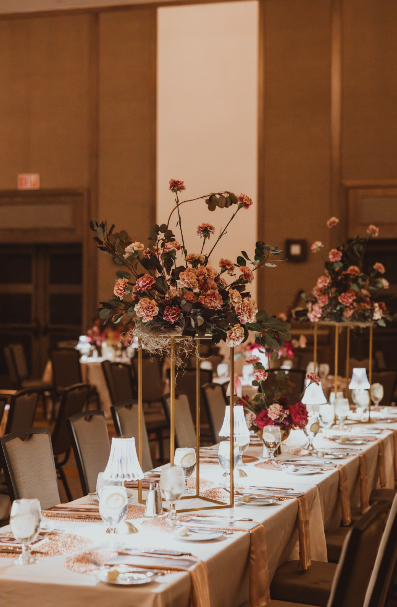Tall flower arrangements in warm terracotta and rust tones on gold stands along a formal reception table with copper chargers.