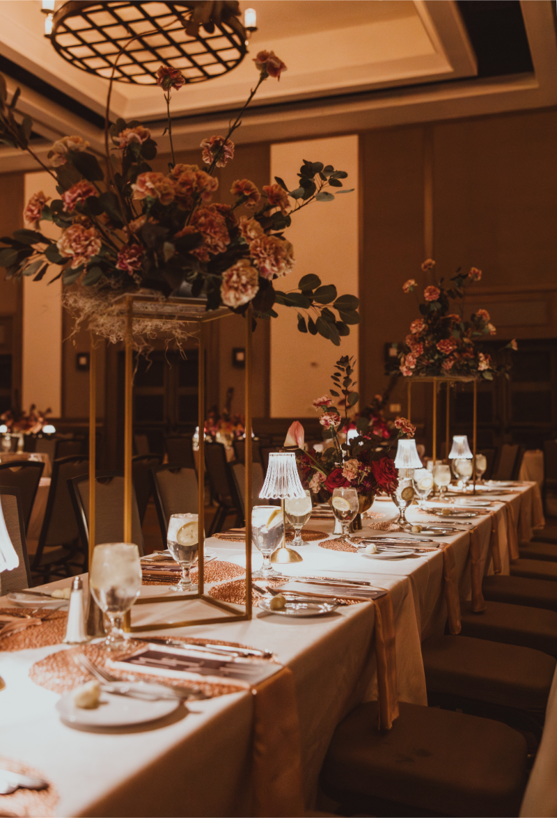 Tall flower arrangements with burnt orange carnations and eucalyptus on gold geometric stands lining a long banquet table.