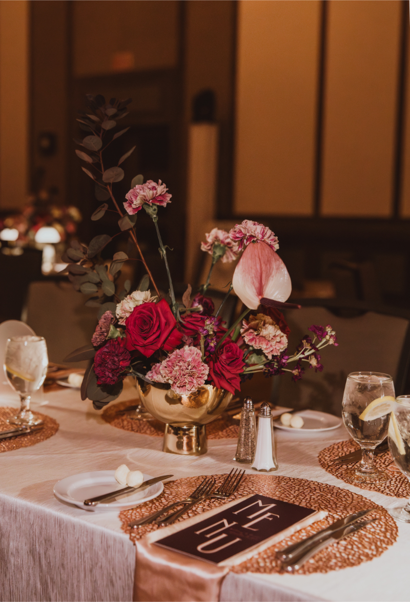 Low gold compote centerpiece with red roses, pink carnations, anthurium, and dark foliage on a round reception table.