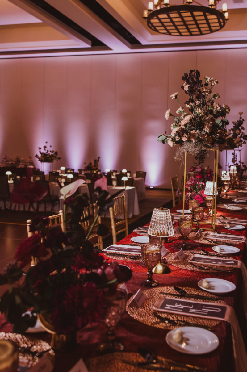 Grand ballroom reception featuring tall flower arrangements and low floral compotes with jewel-toned blooms along a red sequin head table.