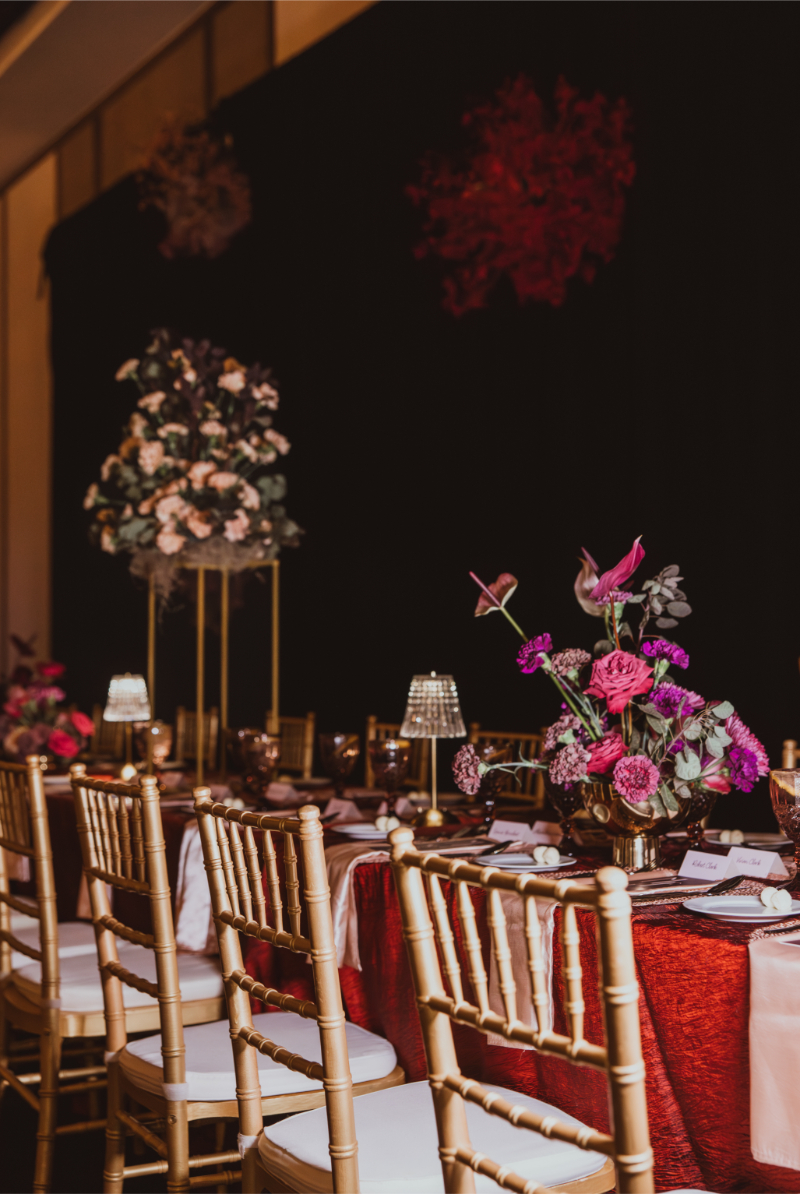 Dramatic reception space with tall flower arrangements, overhead floral orbs in deep red and purple, and gold chiavari chairs.