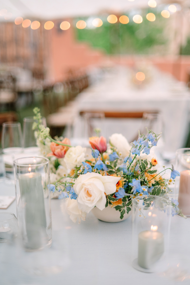 Low centerpiece of white roses, blue delphinium, orange tulips, and wildflowers in a white ceramic bowl on a blue linen reception table.