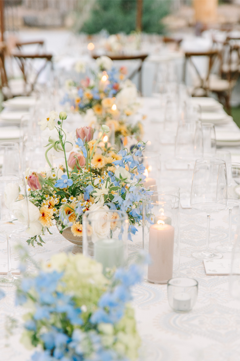 Long reception table with a floral runner of blue delphinium, tulips, marigolds, and white ranunculus with pillar candles at a tented wedding.