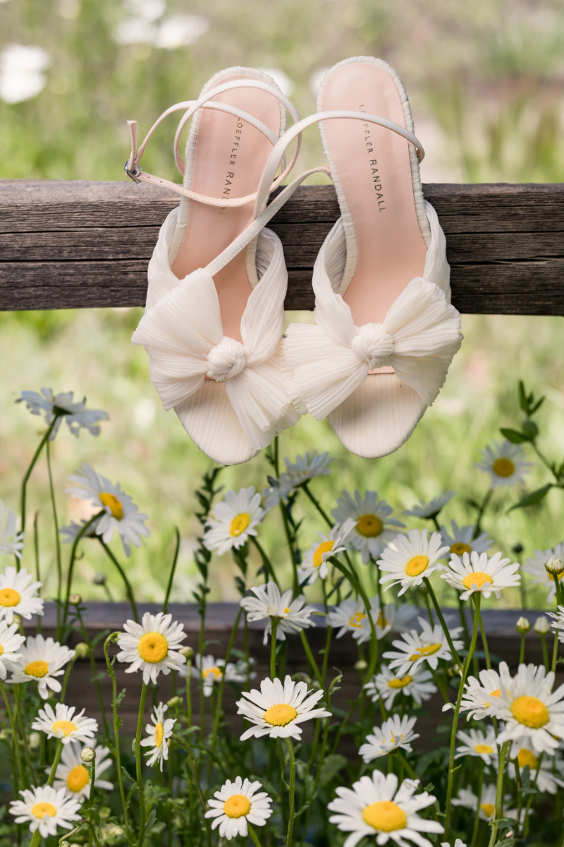 White heels with bow hang from a wooden fence post just above blooming daisies.