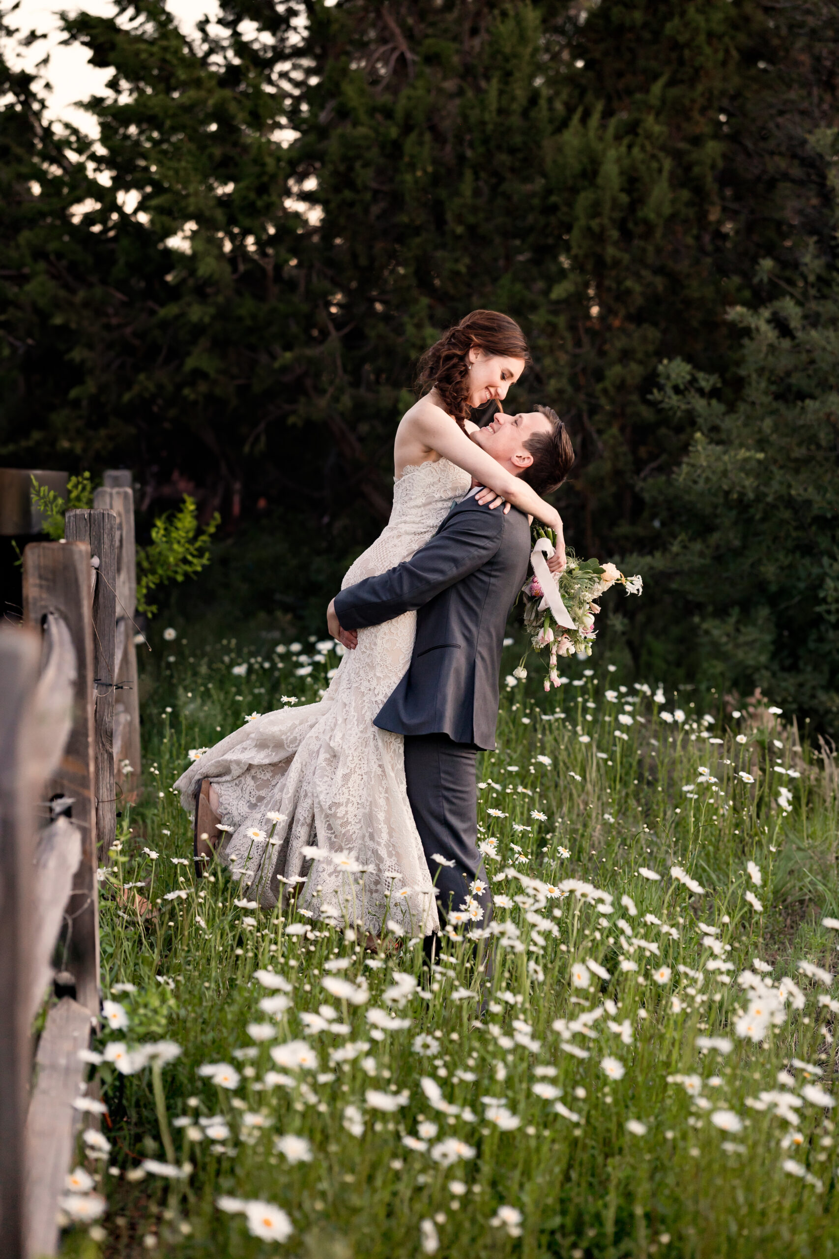 Groom lifts bride into the air in a field of daisies.