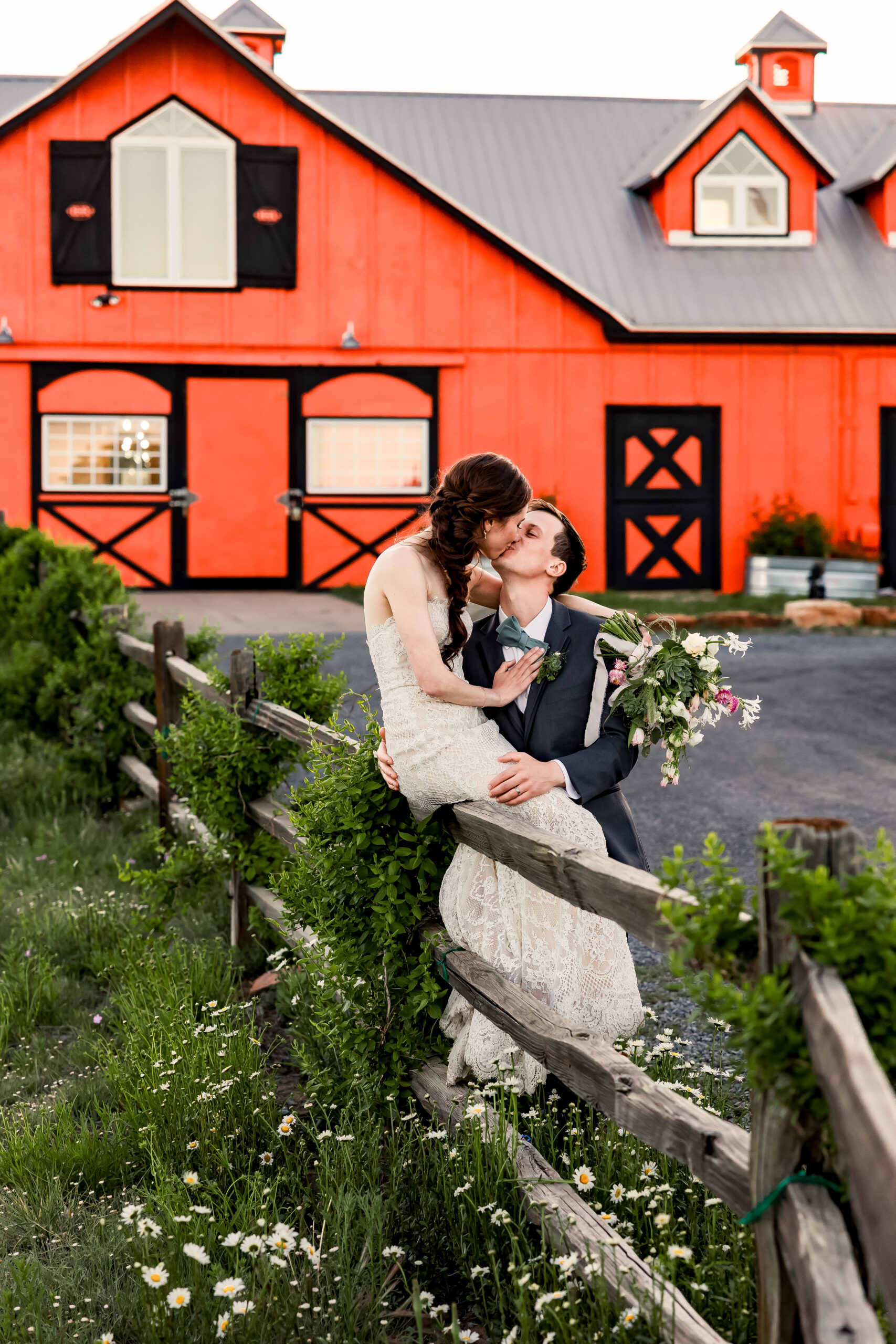 Bride sits on a wooden fence post as she bends down to kiss groom, a red barn wedding venue behind them.