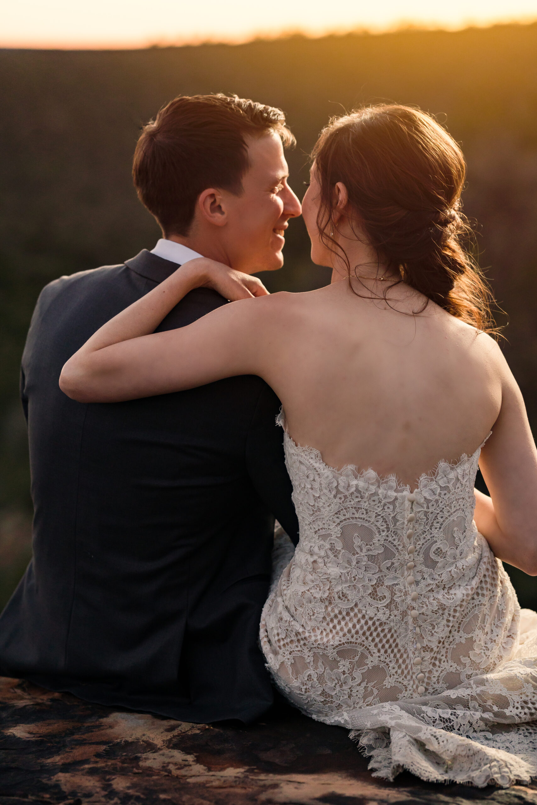 Bride and groom sit on the edge of a cliff overlooking the rolling hills of Tennessee as the watch the sun set.
