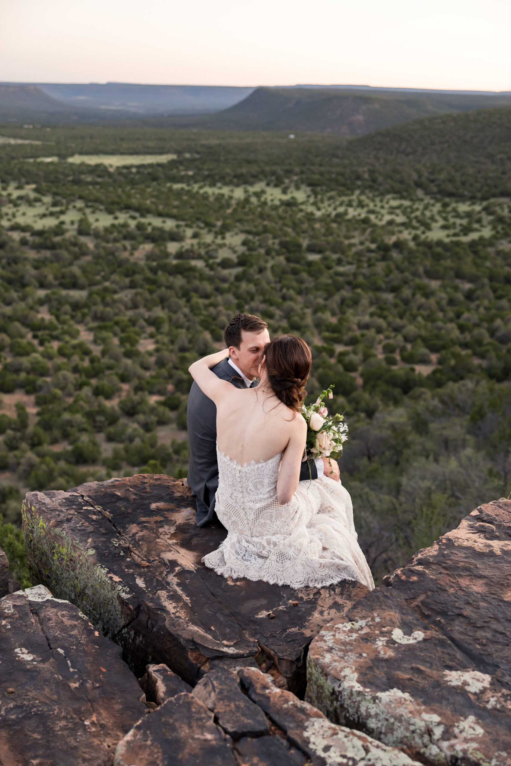 Bride and groom sit on the edge of a cliff overlooking the rolling hills of Tennessee as the watch the sun set.