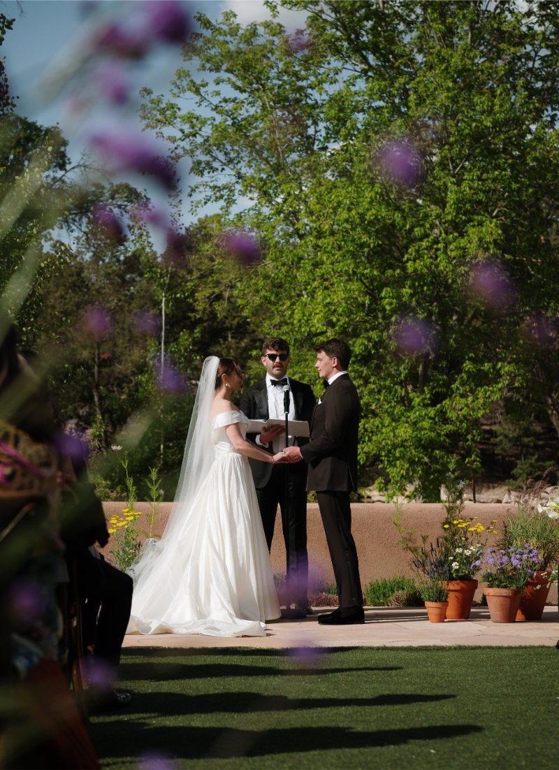 Bride and groom hold hands at the altar with fall wedding flowers blurred in the foreground of the photo, framing the couple.
