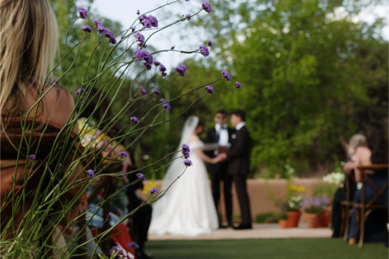 Bride and groom hold hands at the altar with fall wedding flowers blurred in the foreground of the photo, framing the couple.