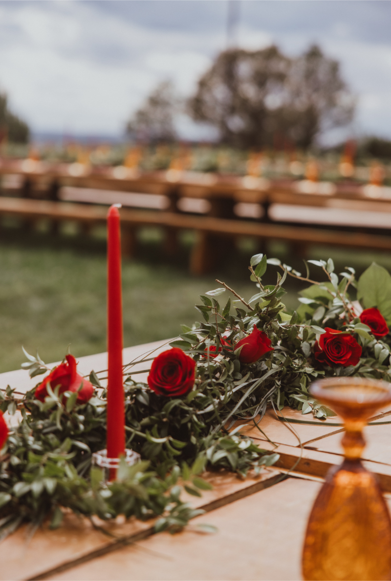 Red pillar candle sitting in glass holder, next to greenery and red florals lined along the center of a reception table.