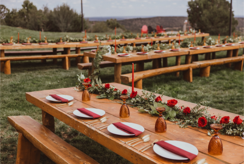 Natural wood reception tables decorated with Christmas wedding ideas featuring red napkins, table candles, greenery, and amber glasses.