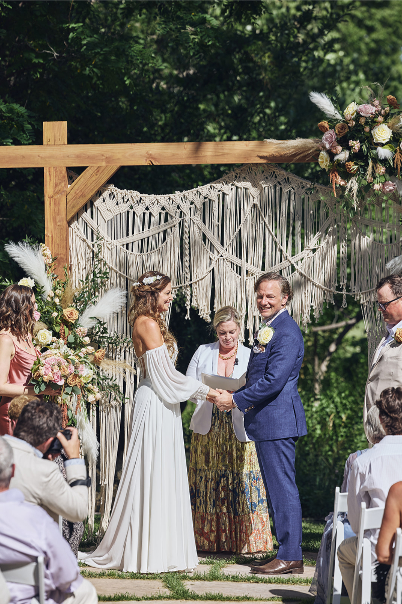 Bride and groom stand under boho wedding arch as they exchange vows.