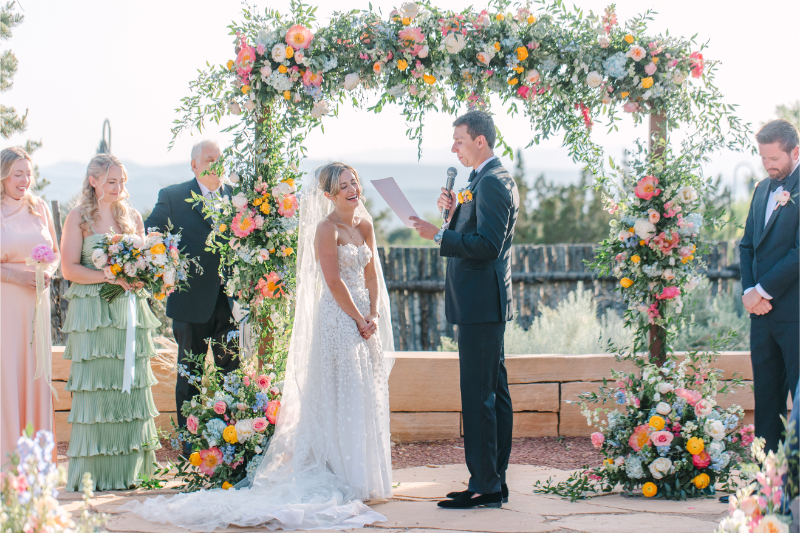 Bride gushes as her groom reads his vows from a piece of paper, standing under the color floral wedding arch ideas the bride always dreamed of.