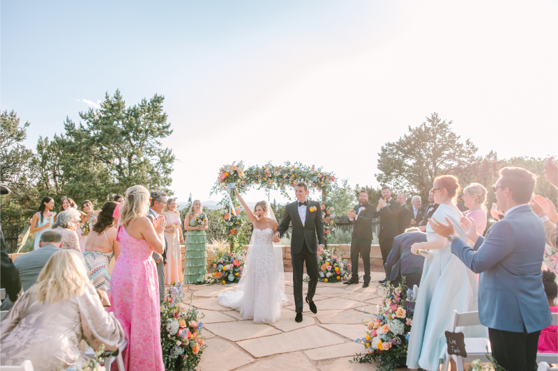 Bride and groom celebrate as they walk down the aisle together after exchanging vows, their colorful wedding arch ideas in the background behind them.