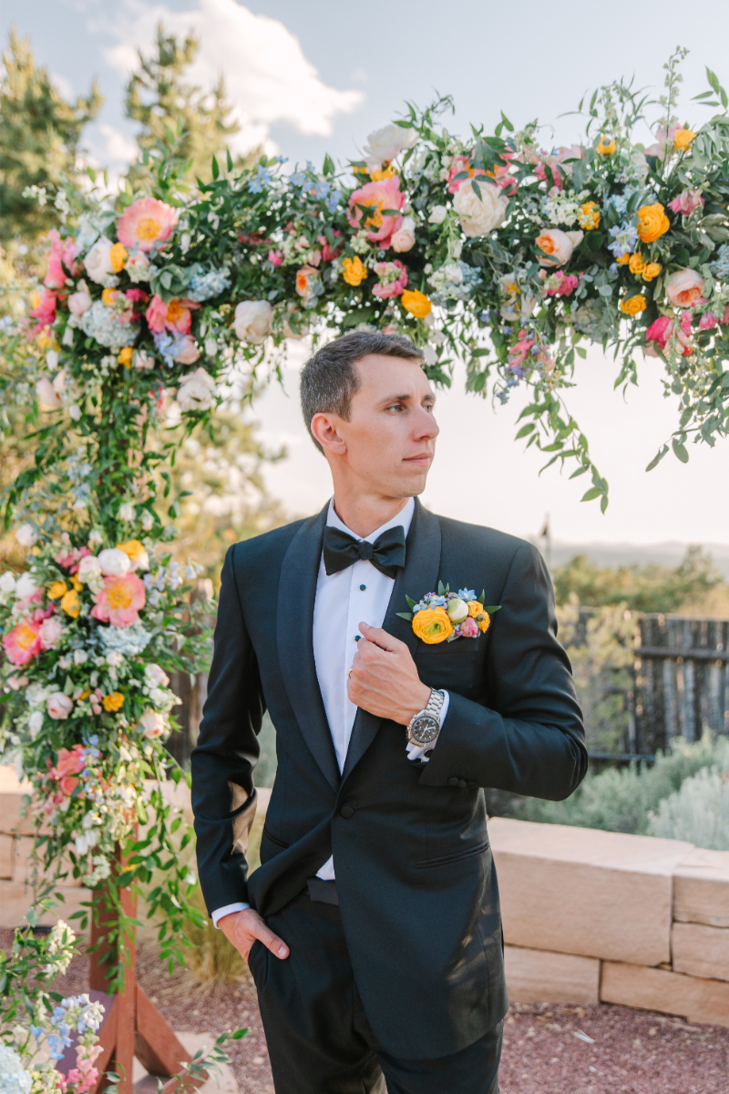 Groom holds onto suit jacket as he poses under wedding arch for a portrait.