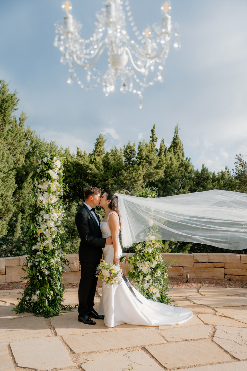 Bride and groom kiss, the bride's veil blowing in the wind behind her, as they pose next to two tall floral installations they planned to use as their wedding arch ideas.
