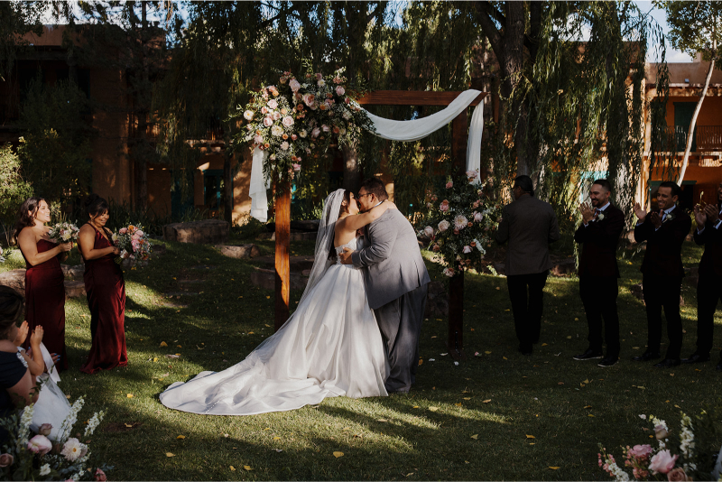 Bride and groom share first kiss under a square wedding arch decorated with florals and draping.