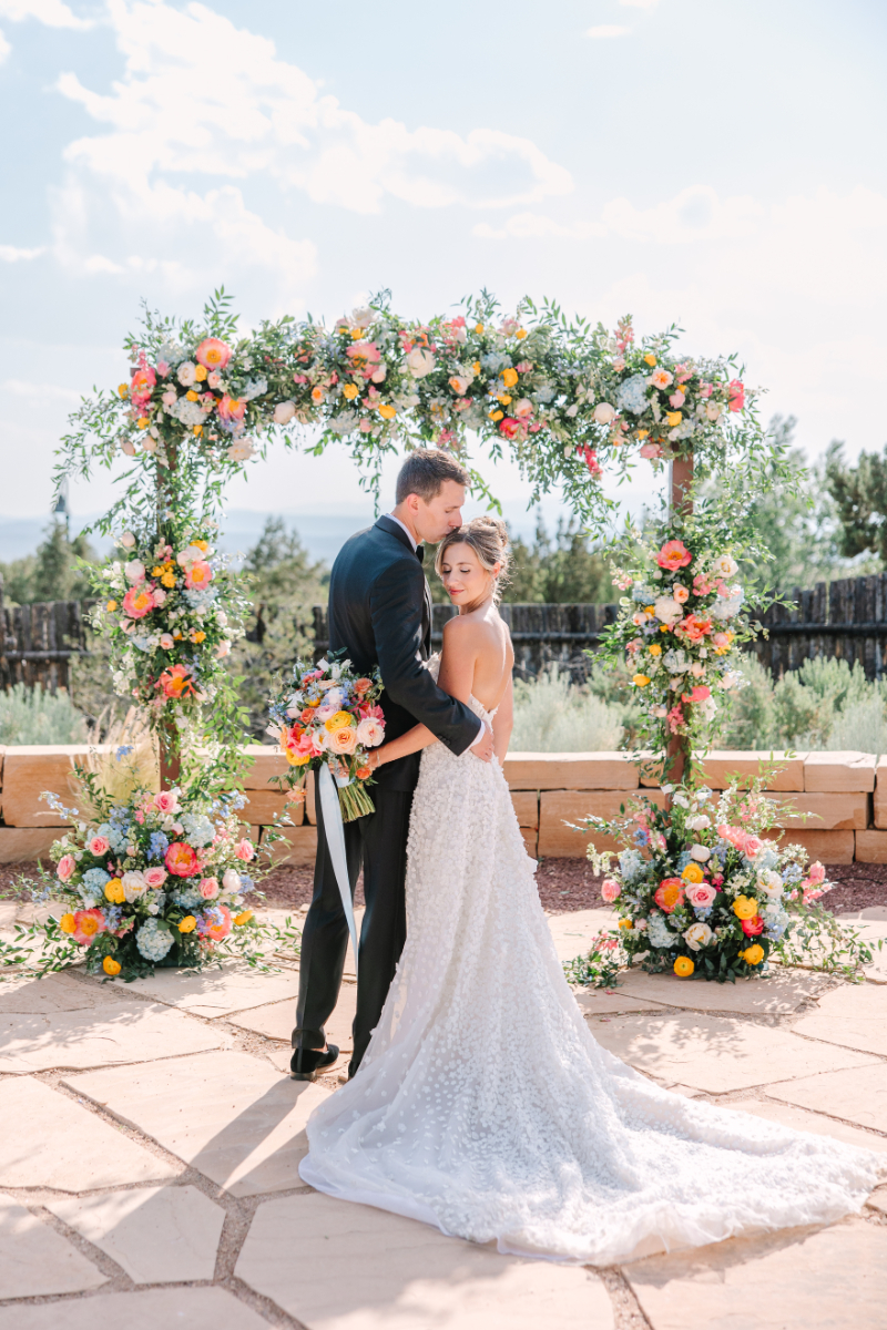 Groom kisses bride on the head as she holds a bouquet that is perfectly cohesive with the wedding arch ideas she had for her ceremony.