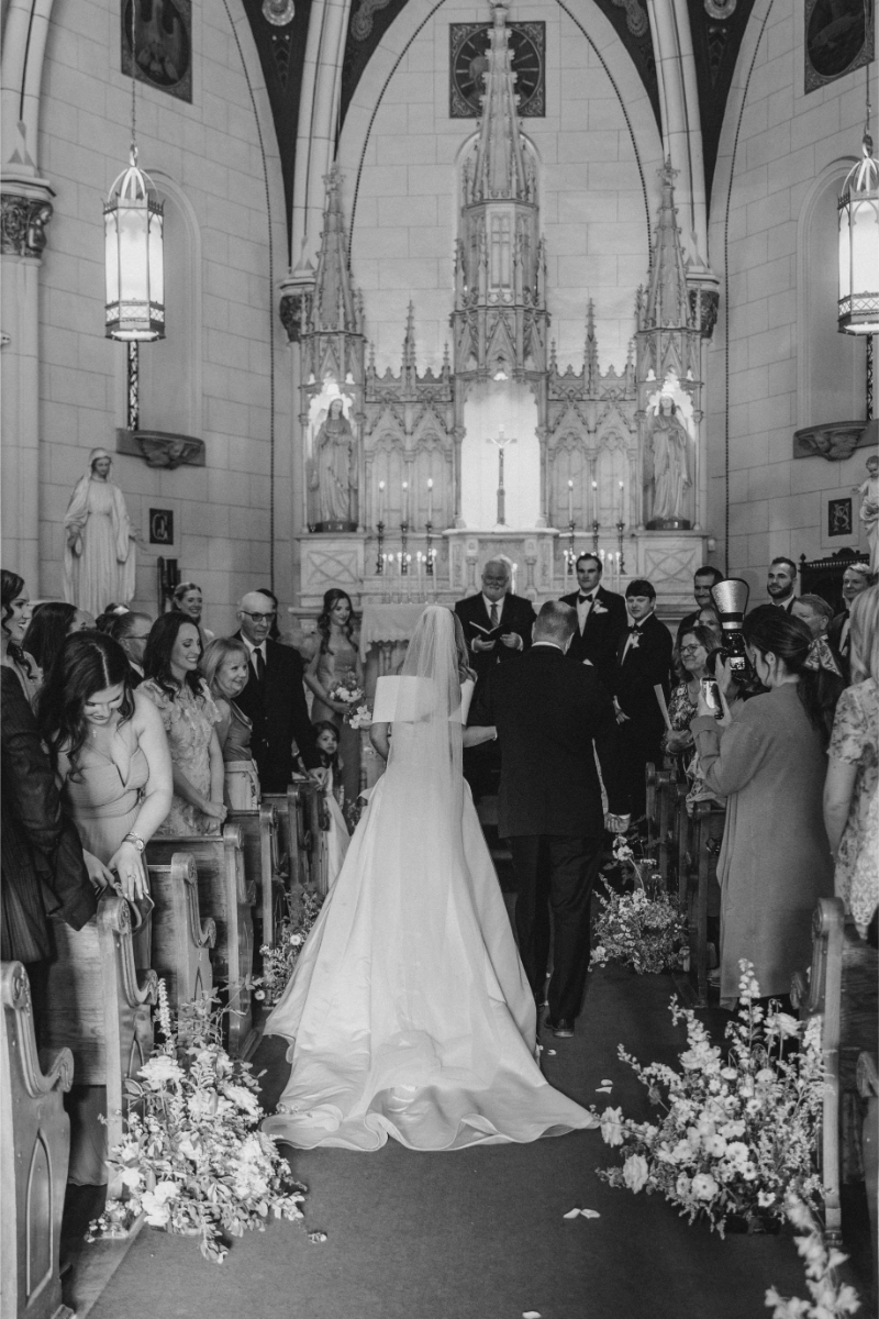 Black and white photo of bride being escorted down the aisle by her father.