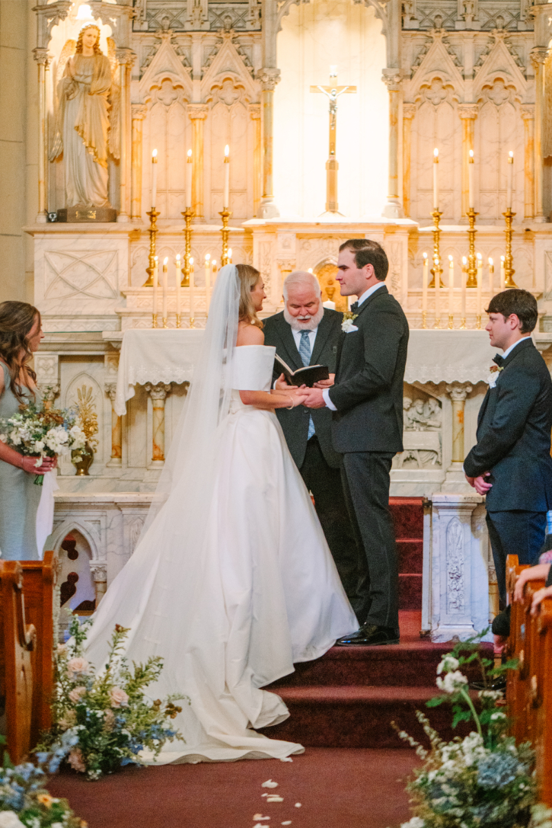 Bride and groom hold hands at the altar as pastor reads from a book.